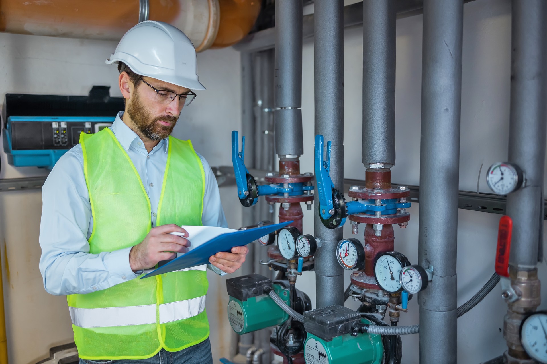 Industrial engineer standing by pipes in heating power plant and make notes in clipboard